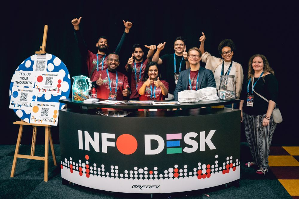 A diverse group of 8 volunteers standing behind the InfoDesk at Øredev 2024, with their hands in the air giving a thumbs up.