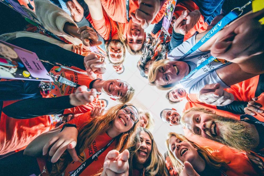 Group photo of the Øredev volunteers, a diverse group wearing orange t-shirts forming a circle and pointing towards the camera in a lively team moment.