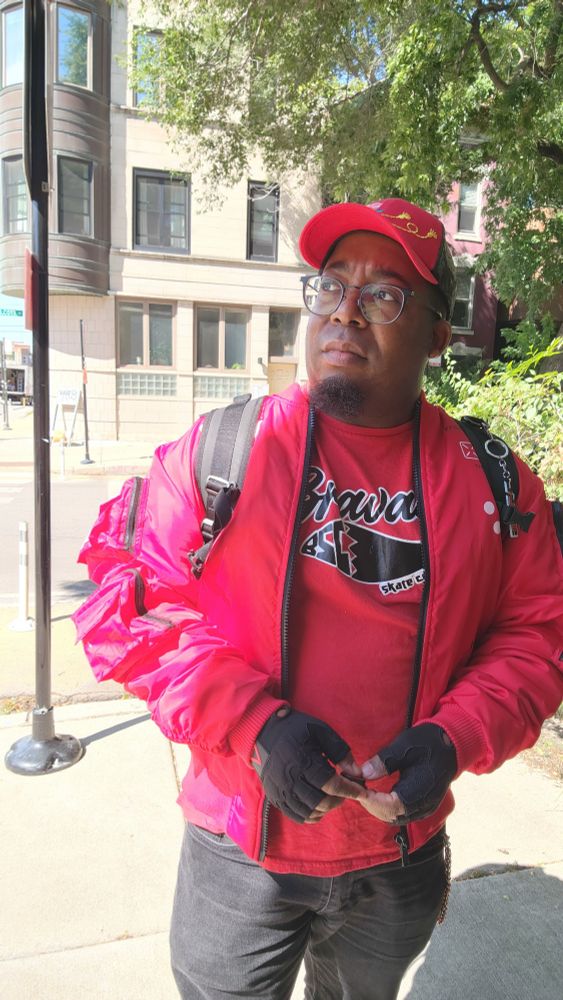 Self shot photo of me, an Afro Latino man, wearing a red jacket, red t-shirt, black jeans, black fingerless gloves, and a red and black hat, in Chicago during the day.