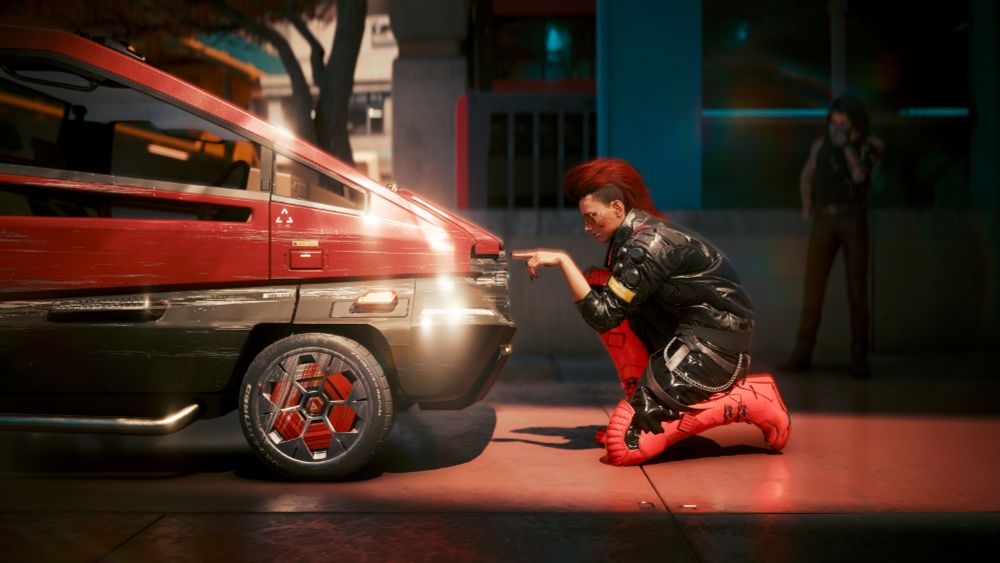V dressed in black and red, with a large bright red mohawk hairstyle, kneels on the pavement of Night City, pointing her finger out as if stopping an also black and red Mai Mai car. Johnny Silverhand stands with his face in his palm in the background.
