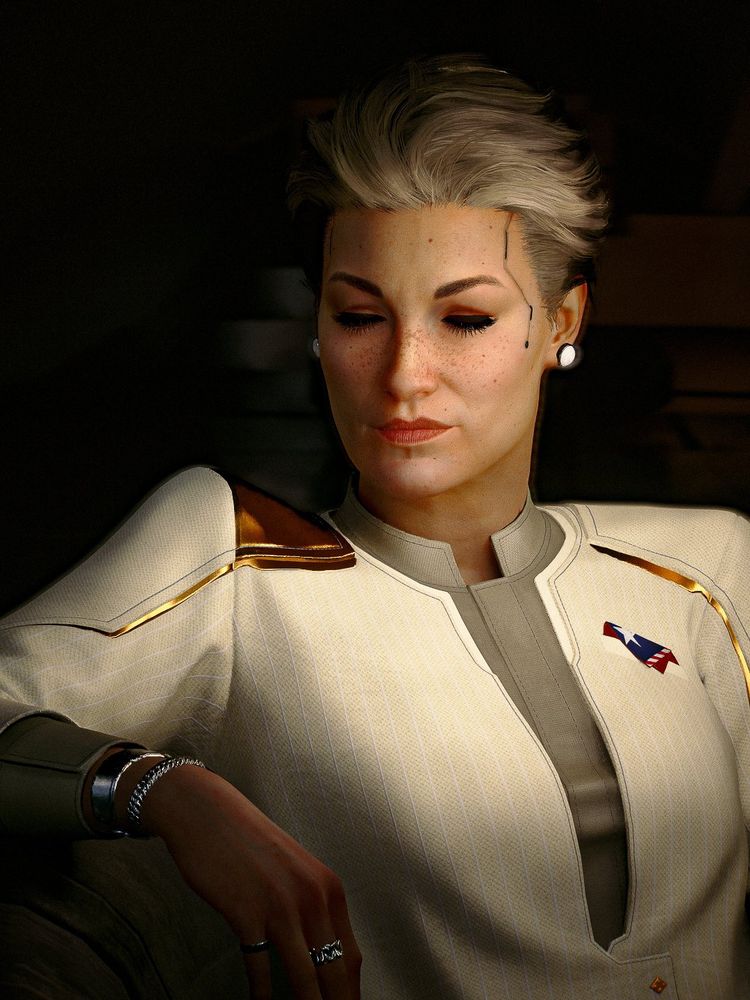 A moody portrait of President Rosalind Myers. Her eyes are downcast and she gives a small smile, freckles covering her face. Her short gray hair is brushed back and gold accents gleam on her white pantsuit.