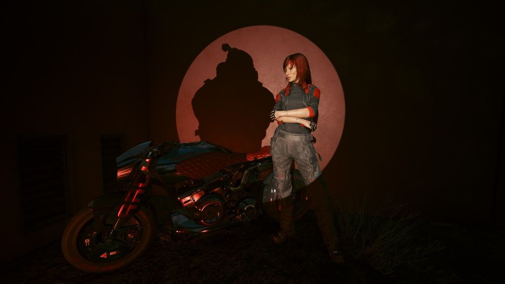 A spotlight is focused on V as she stands next to a red and black motorcycle, mostly in shadow. V wears dark grey, red, and black clothes and stands posing with her arms folded across her front. Her shadow doesn’t belong to her, however, but instead Jackie Welles. 