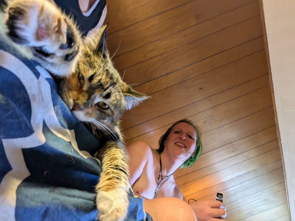 A picture taken from below of a tabby cat lounging with her paws hanging down toward the camera, and my wife leaning back with green hair and a cute smile