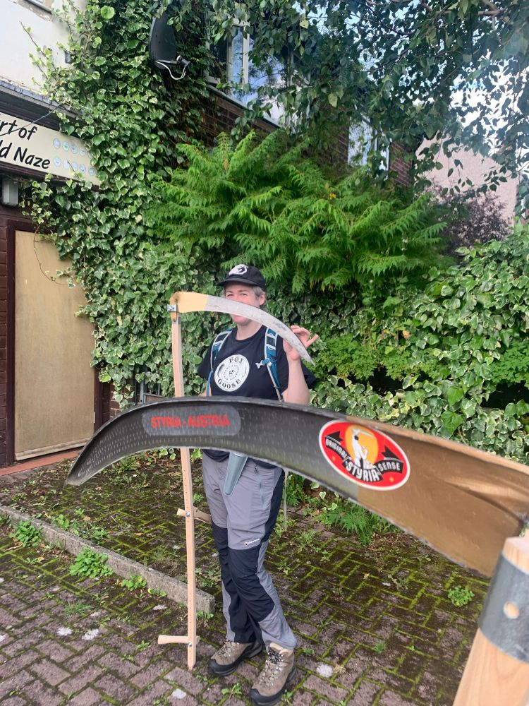 Beck holding and standing behind a scythe. There is a scythe blade in the foreground