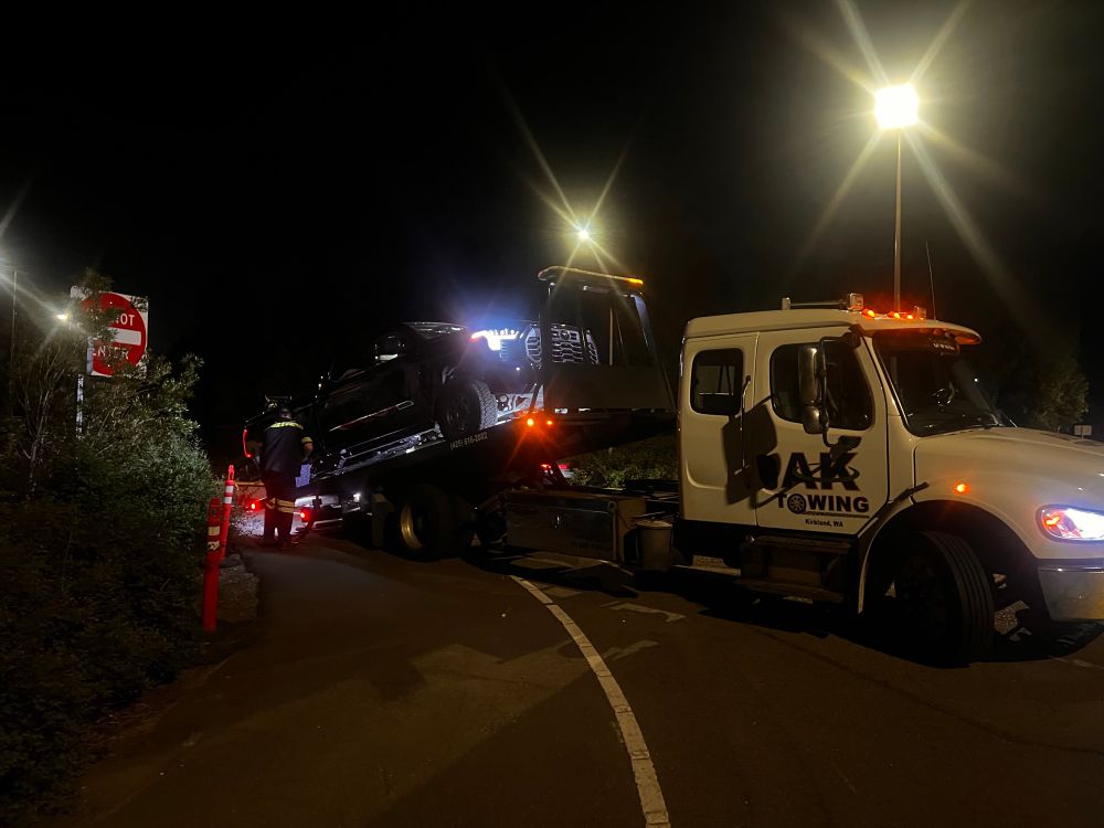 Night scene, with a large tow truck vehicle, and on a slanted bed, they are pulling up a full size truck onto the tow bed from alongside the road by a do not enter sign.