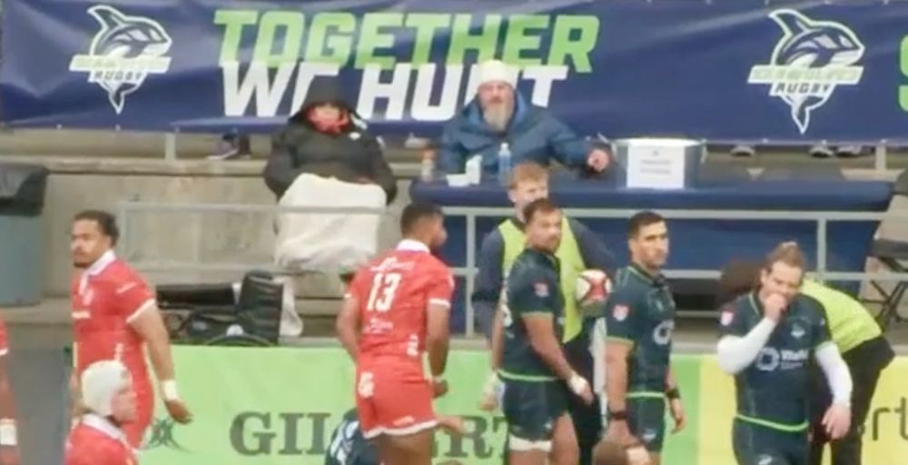 Screen grab from a TV broadcast for a rugby game. Players in red or blue uniforms are in the foreground, background is a banner with Seattle Seawolves blue logo and text: together we hunt. Two fans sit facing the players bundled in cold weather clothing.