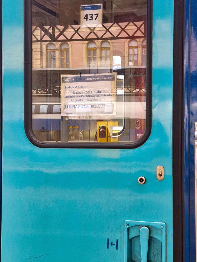 Photo of a train door, blue greenish, with in the window buildings reflected. In the window cards with the car number 437 and destination (Budapest to Cluj-Napoca) 