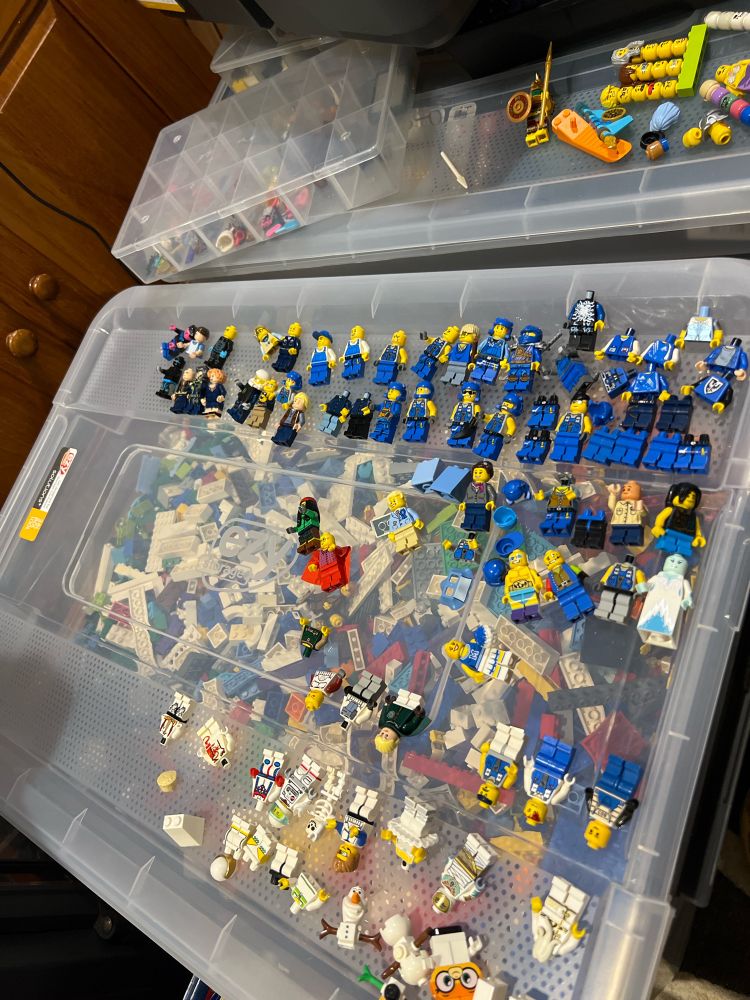 A plastic container full of LEGO, and on top there’s a row of blue minifigures and a row or two of white. In various states of completion. 