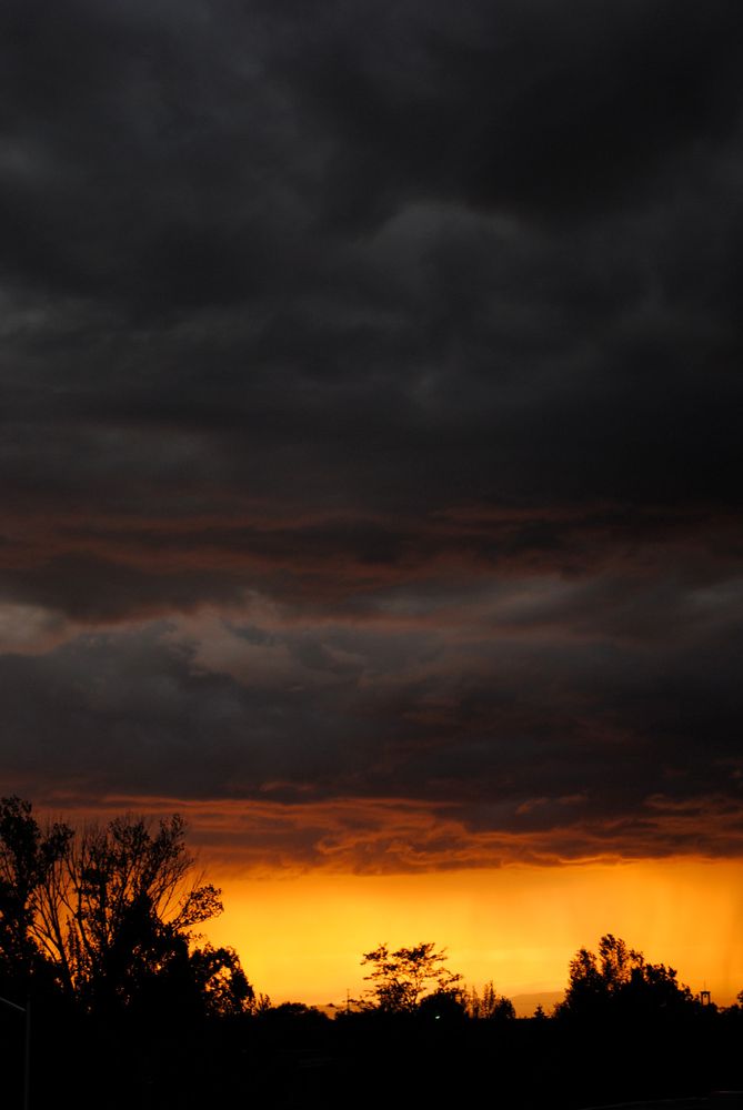 Massive gray and black thunderclouds fill most of the photo. At the bottom is a brilliant slice of orange from the setting sun. 