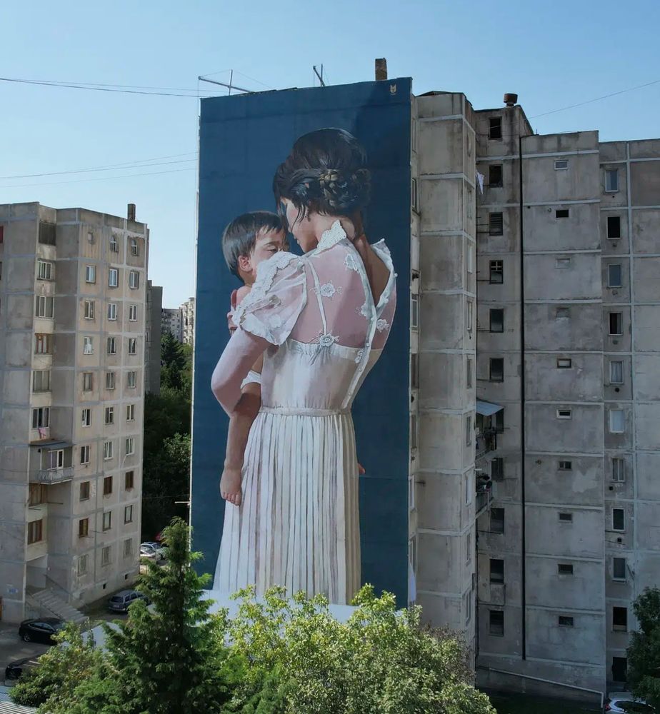 Street art mural of a mother holding her young child. The mural is on an eleven story building in the country of Georgia. The mother wears a delicate cream-colored dress. Child and mother have eyes partially closed with a look of content and peace.