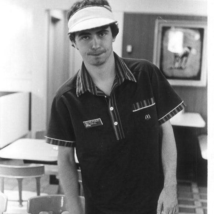 A black and white photograph of a young Daniel Johnston working the day-shift at McDonald's in Austin, Texas. 1984. He wears a McDonald's uniform and leans slightly to the side while staring directly into the camera. The restaurant is empty.

Photo credit of the musician is unknown. 