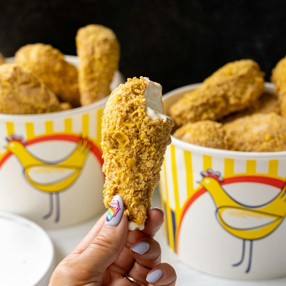 A hand holds an ice cream treat disguised as a fried chicken drumstick. One bite is missing, revealing the vanilla ice cream within. In the background are two colorful paper buckets in white, yellow and red. The buckets are filled with more ice cream treats disguised as fried chicken