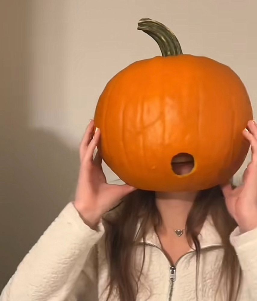 Woman with a pumpkin on here head with a hole where her mouth is. 