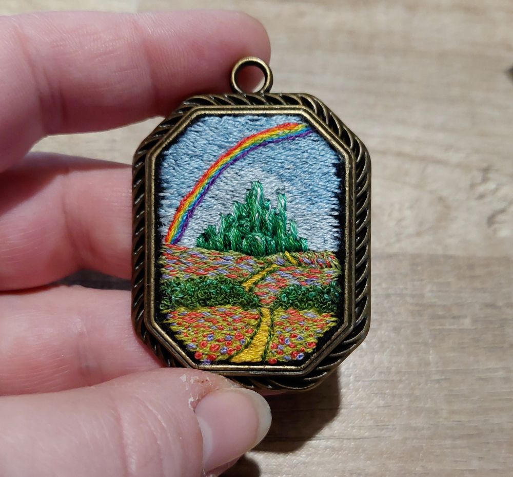 Pendentif octogonale en longueur. C'est un médaillon. Dessus il y a une broderie d'un paysage. C'est le pays d'Oz. On voit la route de briques jaunes, la cité d'émeraude et un arc-en-ciel.