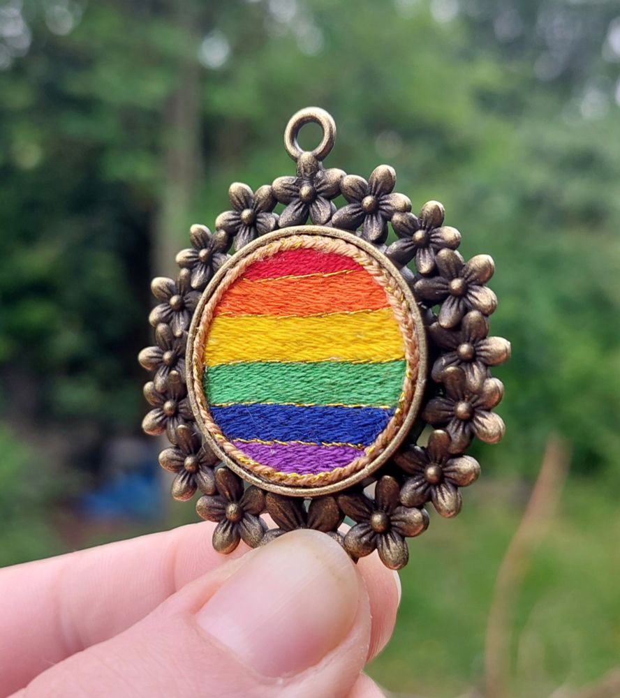 Photo qui montre un pendentif en métal avec une couronne de fleurs. Le centre est une broderie ronde avec des rayures horizontales rouge, orange, jaune, vert, bleu, violet. Il y a une séparation en fil d'or, ainsi qu'une couronne de fil d'or tressé tout autour.