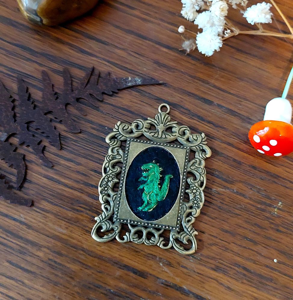 Pendentif en forme de cadre ouvragé. Au centre il y a une broderie ovale d'un dinosaure sur fond noir.
Le pendentif est posé sur une surface en bois.