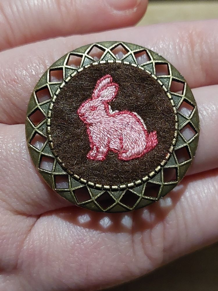 Broche ronde en métal à motif rosace. Au centre une broderie sur feutre marron. La broderie est un lapin rose.