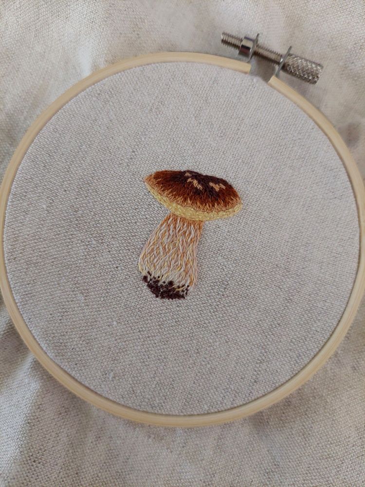 Photo d'une broderie. Le tissu est écru et fixé sur un tambour à broderie. La broderie est un cèpe.