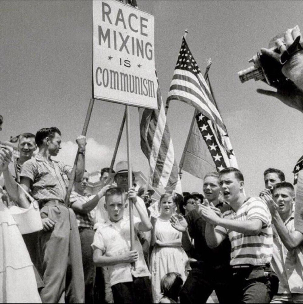 a black and white photo from the 1950s of a crowd with prominent American flags and a sign that reads:

RACE MIXING IS COMMUNISM