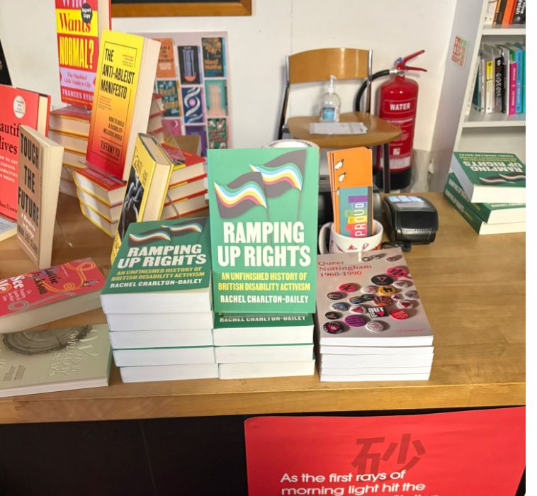a stack of my book ramping up rights which is green with a disability pride flag on the front and the subtitle an unfinished history of british disability activism in yellow then my name in white. just behind my book and to the side is a selection of other disabled books