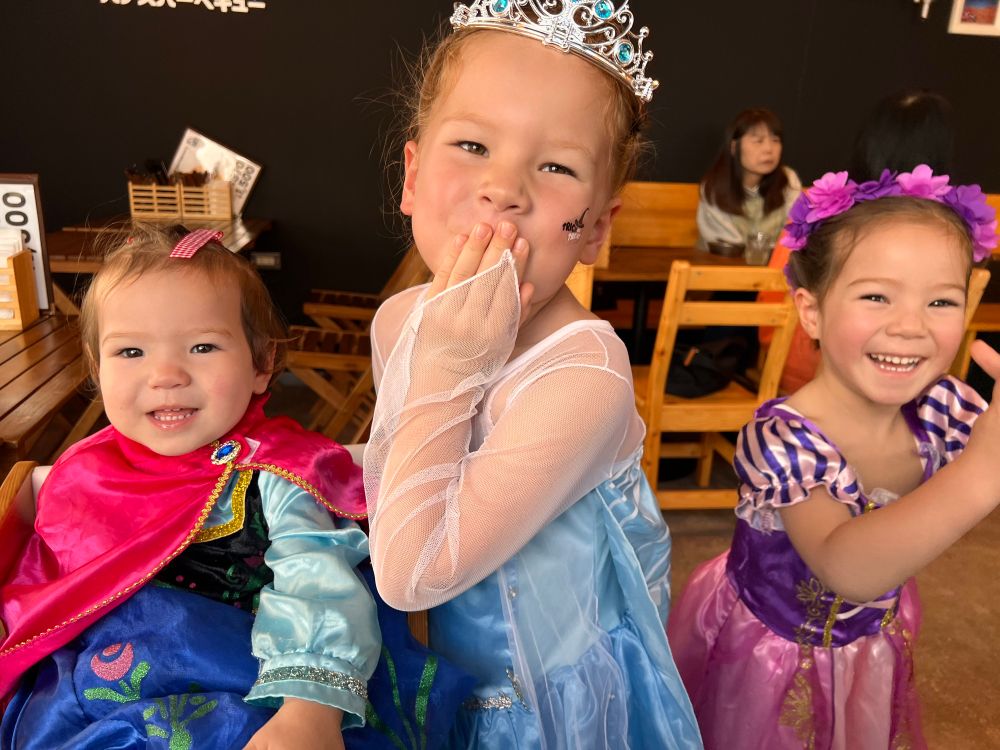 A 1, 5, and three year old, dressed in princess costumes, smiling and blowing kisses 