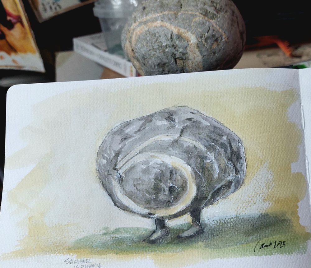 Cuaderno von una pintura a témpera de un canto rodado con piernecitas. Es gris, con una espiral de cuarzo blanco.
Al fondo de la foto se ve la piedra real.