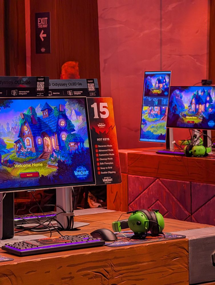 A wide-angle photo of a World of Warcraft "Housing" demo station at Gamescom. The setup features two large vertical monitors showing a detailed and vibrant in-game house with a "Welcome Home" message. A keyboard with purple backlighting and a green, goblin-themed headset with a microphone and a speaker are visible on the wooden table. A sign with the number "15" and a "Hot Keys" list is visible between the two monitors. Other gaming stations with monitors are visible in the background, along with a red-lit "EXIT THIS WAY" sign. The booth is branded with the World of Warcraft logo and promotional material for the game's new housing feature.