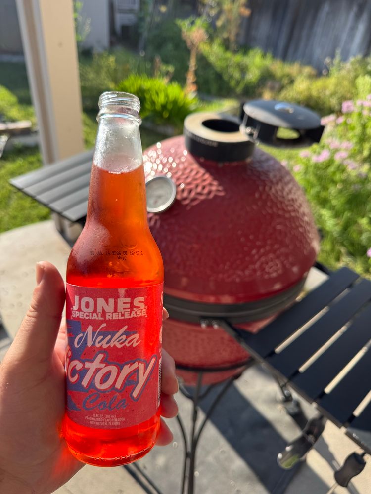 A hand holding a translucent pink Jones Special Release Nuka Victory soda with a grill, patio and garden out of focus in the background.