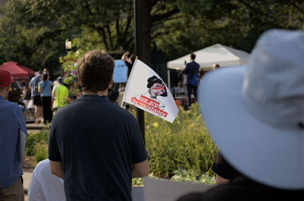 Peaceful protesters stand in the blazing sun. A man holding a small flag that reads "We are the Resistance" under a stylized image of Carrie Fisher as Princess Leia from the Star Wars franchise