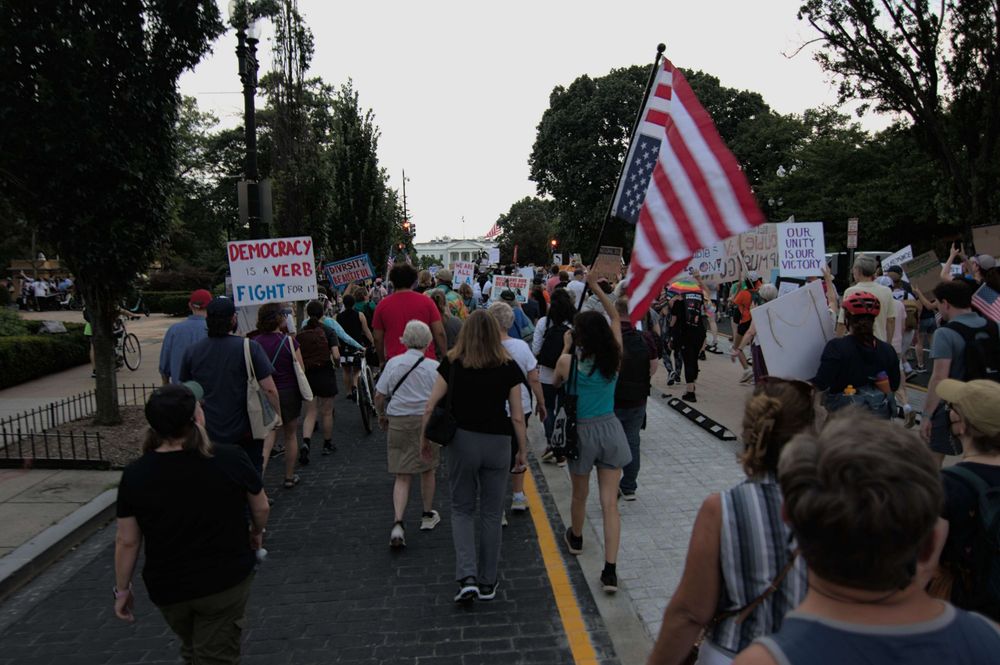 Many peaceful protesters march with the White House in the background. Signs read "Democracy is a Verb, Fight for It" "Diversity is Beautiful" and "Our Unity is Our Victory" a woman marches with a large United States Flag, flown upside down, denoting a sign of distress