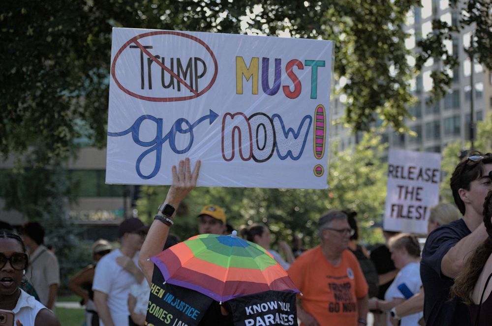 Many peaceful protesters assemble in the shade of the park trees. One wears a rainbow parasol hat and holds a sign reading "Trump must go now" in stylized fonts