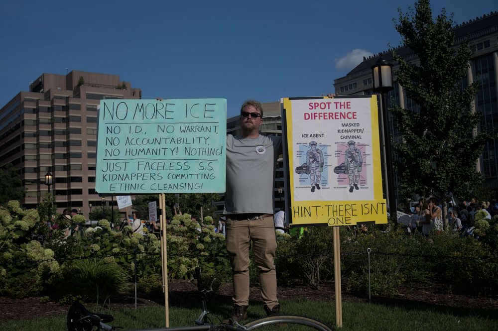 A gentleman poses with two signs. The one on the left reads "No More Ice; No I.D., No Warrant, No Accountability, No Humanity, Nothing!; Just Faceless S.S. Kidnappers Committing Ethnic Cleansing" and the other reads "Spot the Difference: ICE Agent; Masked Kidnapper/Criminal" where two identical images of masked individuals in riot gear stand, it closes "Hint: There Isn't One"
