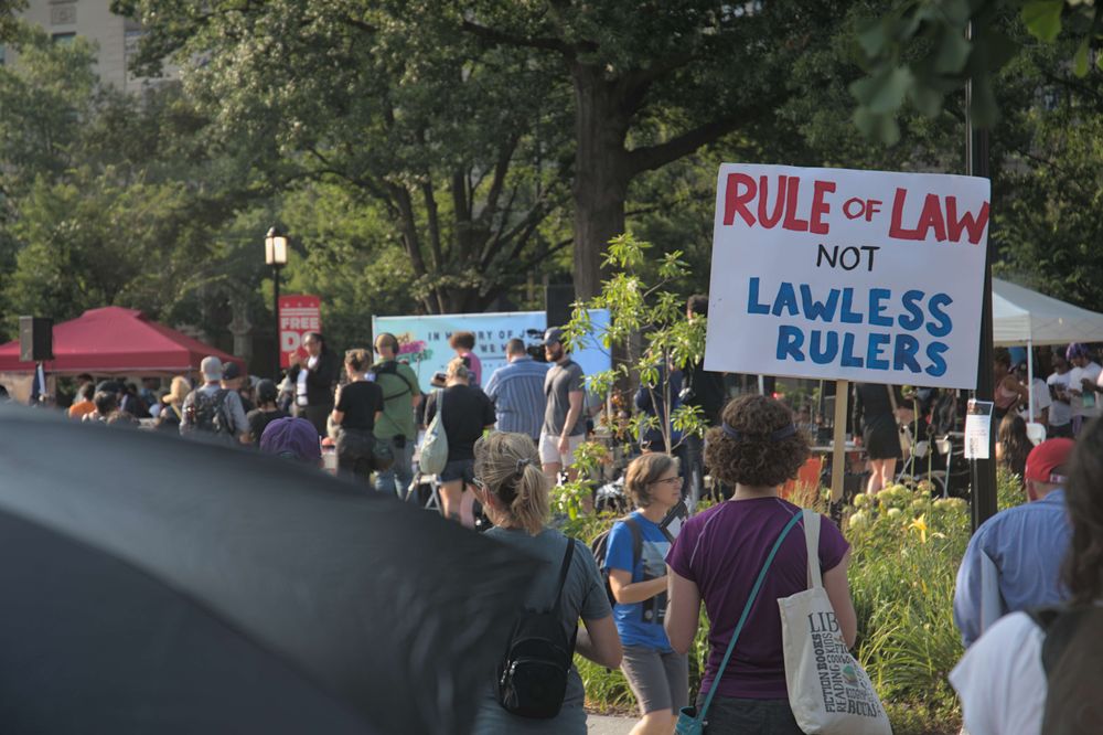 Many peaceful protesters watch as event organizers address the crowd, a protest poster in the foreground reads "Rul of Law not Lawless Rulers"