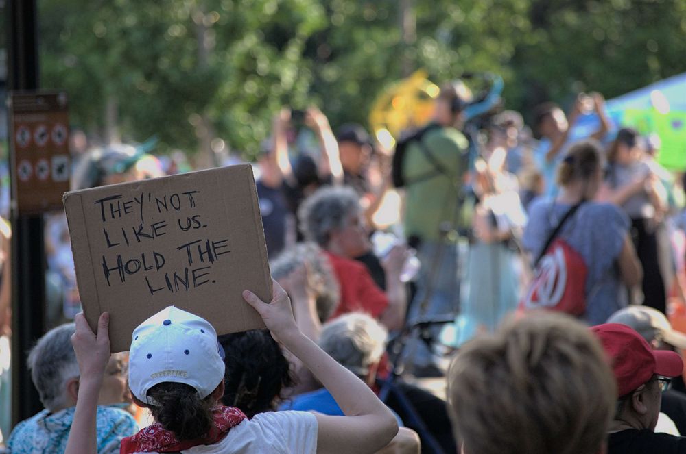 Many peaceful protesters listen to event coordinators while holding signs, one in the foreground reads "They Not Like Us. Hold the Line"