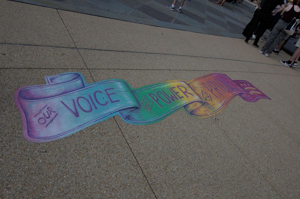 A rainbow banner applique, in the style of chalk art, reads "Our Voice Our Power Our Pride" with the DC flag insignia at the end