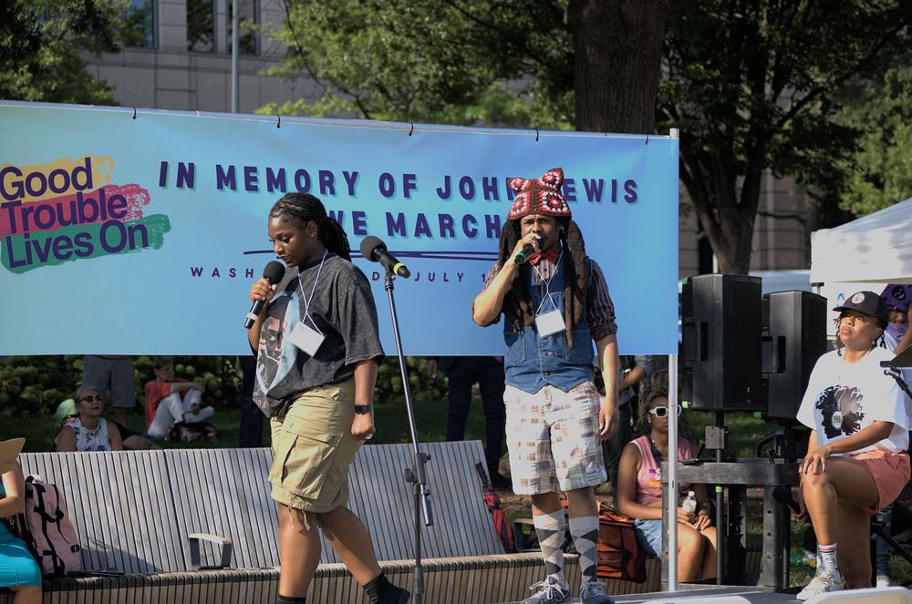 Musical duo, a woman and a man, performing for peaceful protesters on stage in front of a banner reading "Good Trouble Lives On; In memory of John Lewis we March"