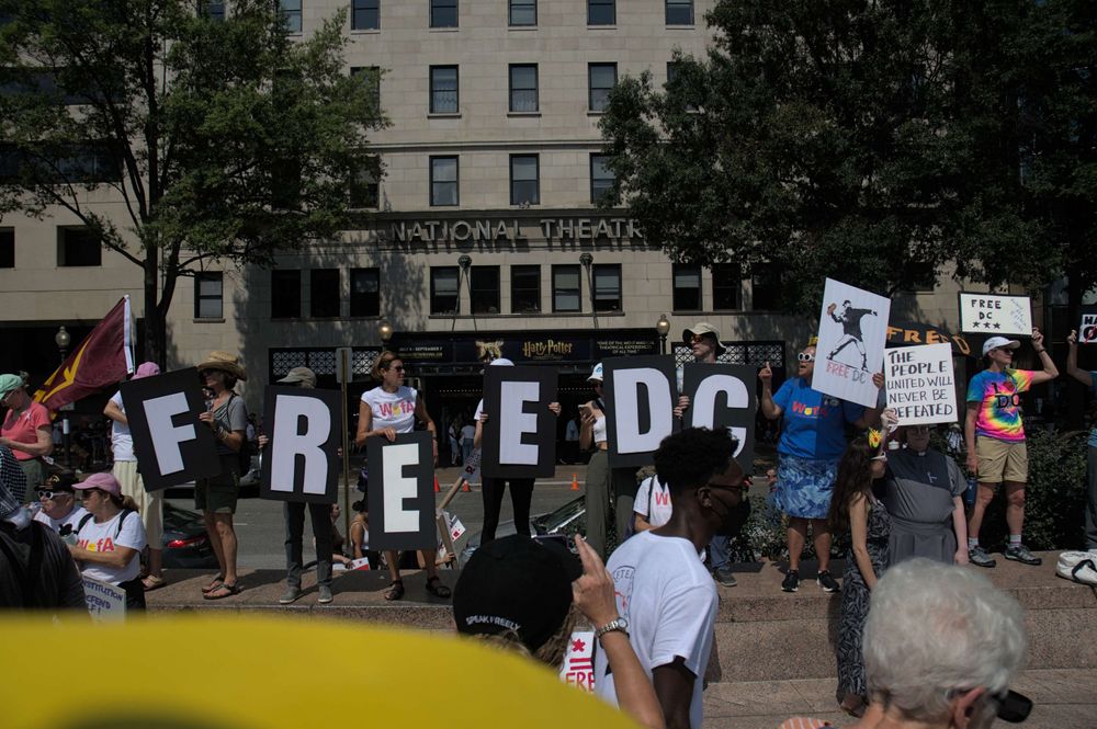 Peaceful protesters at the end point of the march stand in front of the National Theater holding individual signs that spell out FREE DC; A sign to the right of them feature "Sandwich Guy" in the stylized graffiti pose