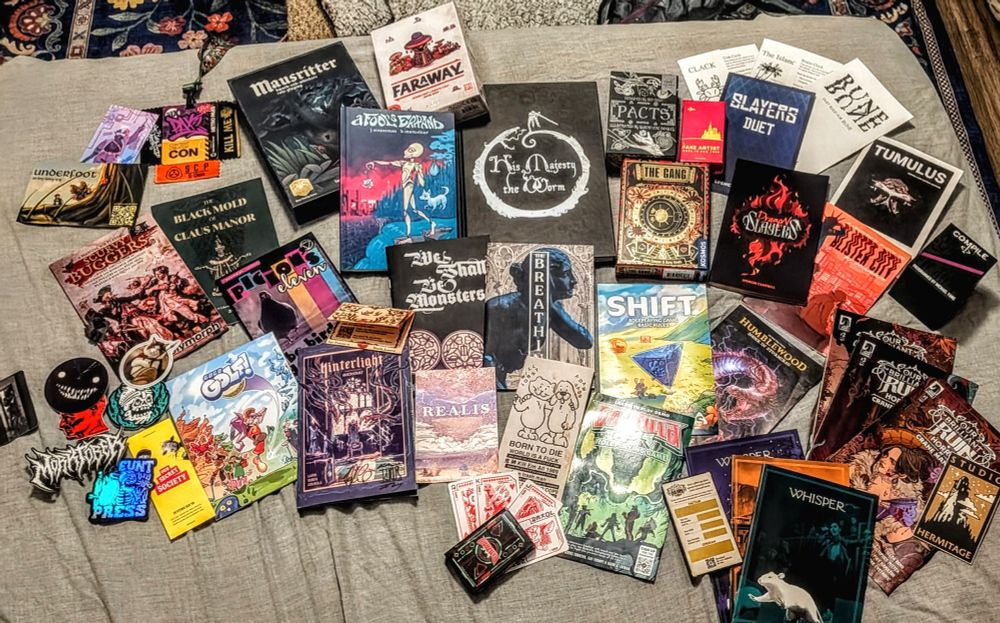 A photo of several tabletop RPGs and board games.

From the top going left to right there is the mausritter box set, far away, a fool's errand, his majesty the worm, pacts, fake artists, slayer's duet, rune duet, tumulus, dragon slayers, blister City, the gang, compile, the black mold of claus Manor, scurvy buggers, pigeons 11, we shall be monsters, breathing, shift, humblewood, our beautiful Ruin, golf, hinterlight, Godzilla rpg, whisper, and several smaller stickers plus a deck of cards.