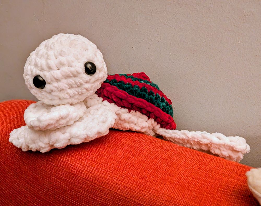 A crocheted white sea turtle with a red and green shell resting its head on its front flippers.