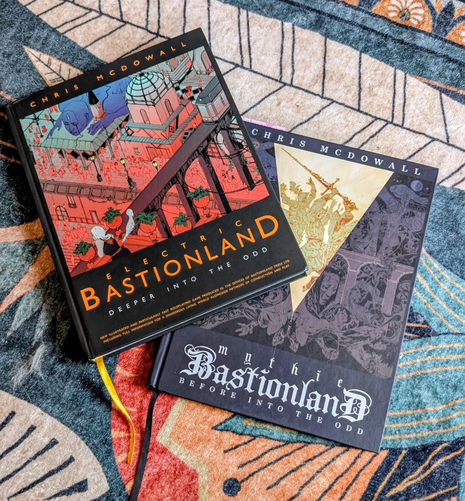 A photo of books, featuring electric bastionland and Mythic Bastionland both by Chris McDowell, on a colorful carpet.