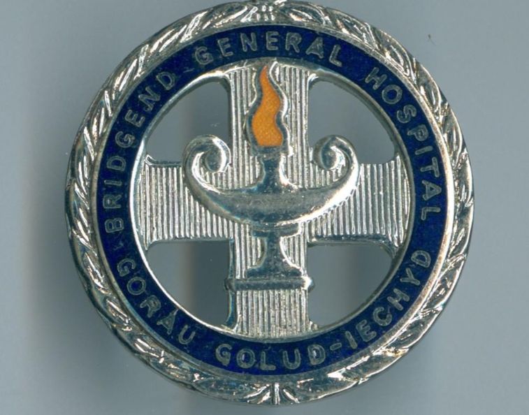 'Silver' circular badge with dark blue enamel.  The outer ring is embellished (possibly a laurel wreath).  Within that is a dark blue enamel ring inscribed ' Bridgend General Hospital, Gorau Golud-Iechyd'.  According to Google Translate the latter is Welsh for 'Best Wealth-Health'.  The centre is a cut out textured cross, at the very centre superimposed is an oil lamp (aka Nightingale lamp) with a yellow enamel flame at its centre.  If anyone knows if the Welsh translation is accurate I'd love to know!
