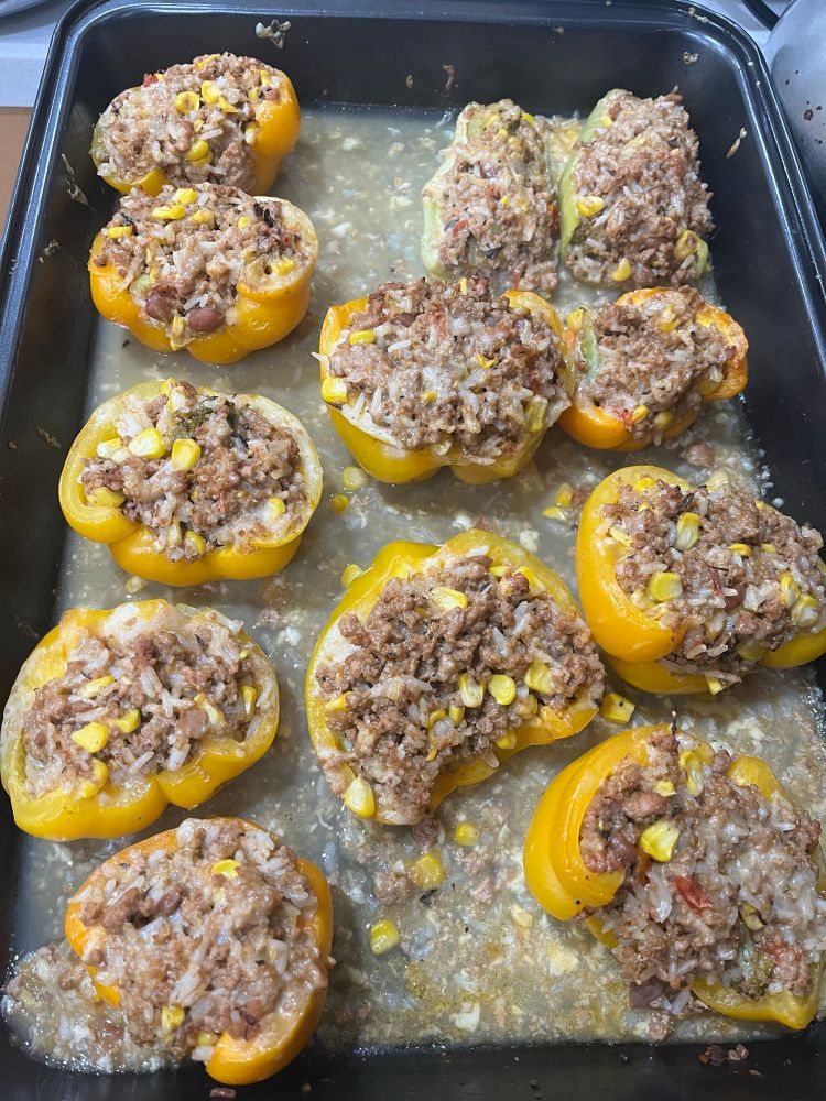 A baking tray filled with various stuffed peppers using that same ground turkey from before but with additional ingredients like corn and Gouda cheese. The bottom of the tray is covered with a chicken stock mix and some fallen off filling. 