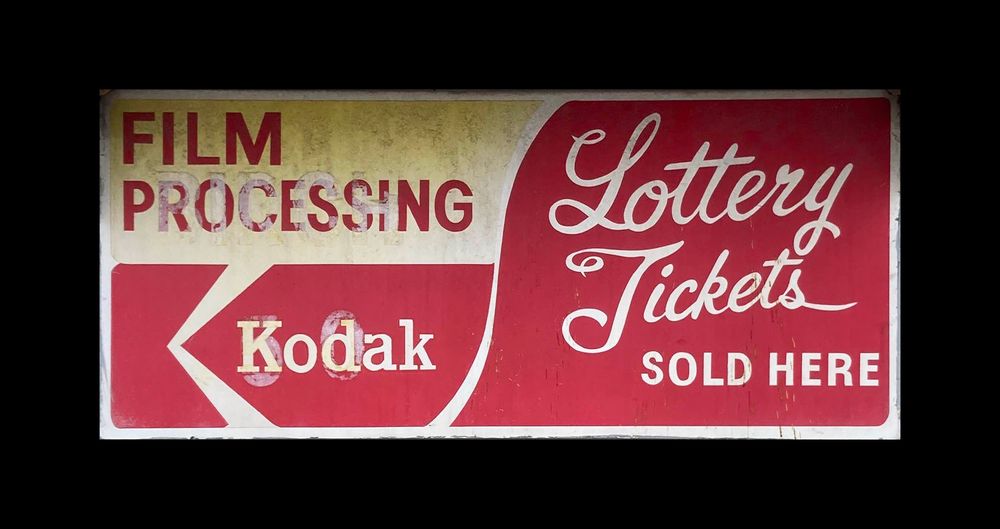 A hand-painted sign, clearly painted on top of a previous sign, on the left it says “FILM PROCESSING” atop the old Kodak logo, then on the right, in script lettering ‘Lottery Tickets’ with a smaller ‘sold here’ underneath.
