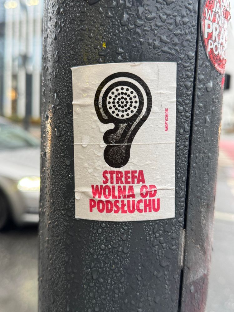 Wlepka na jakimś słupie z ilustracją przestawiającą ludzkie ucho i czerwonym napisem “Strefa wolna od podsłuchu”