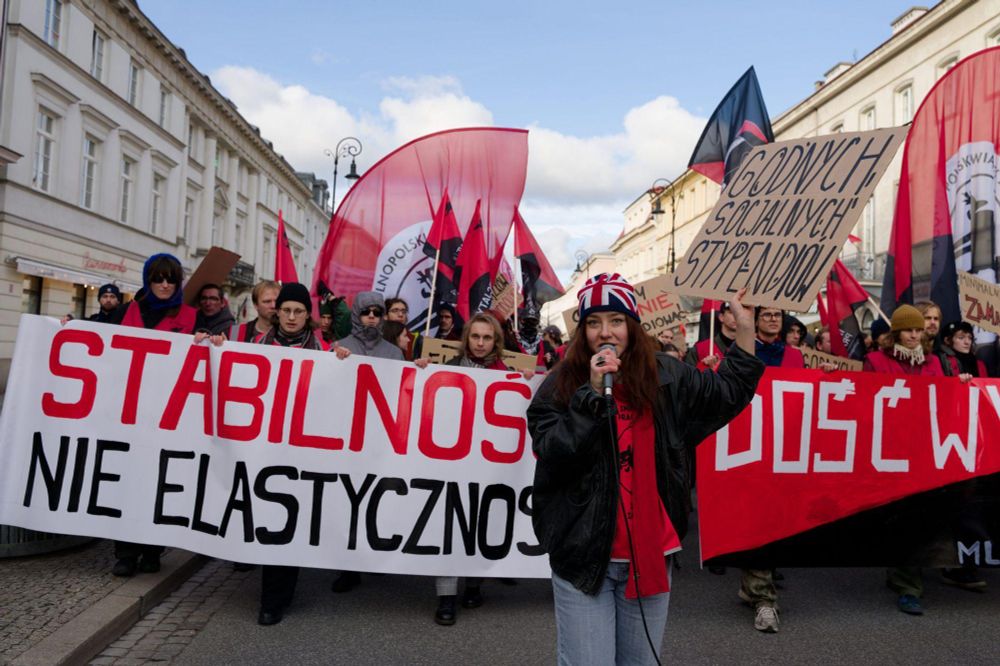 Osoba z transparentem przemawiająca do mikrofonu. Za nią kroczy blok z dwoma banerami. Na jednym jest napisane "stabilność nie elastyczność", na drugim "dość wyzysków młodych".