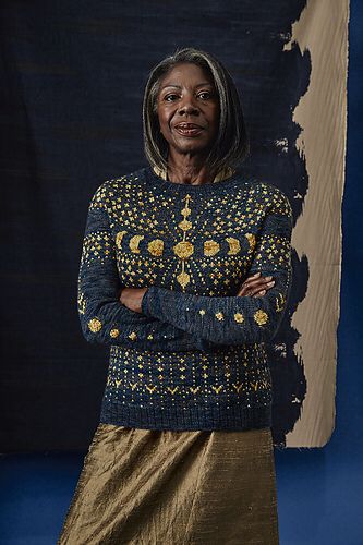 A black older woman wearing a midnight blue knitted sweater. The golden colorwork of stars and the moon phases makes it look like a starry sky. 