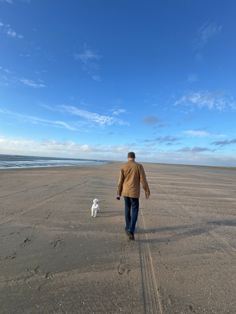 Ein langer breiter Sandstrand nimmt 2/3 des Bildes ein. Rechts das Meer, oben blauer Himmel mit ein paar Wolken im Hintergrund. In der Mitte läuft ein Mann in beigefarbener Jacke und dunkelblauer Jeans, der einen kleinen weißen Hund an der Leine führt. Beide sind von hinten geknipst
