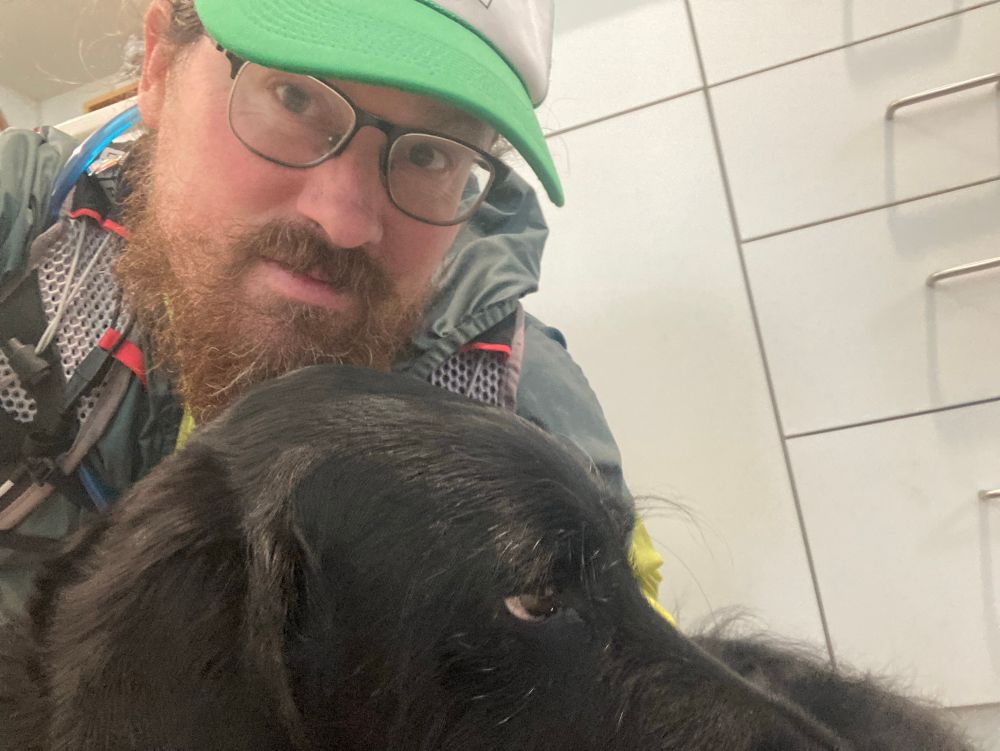 A black dog snuggling into a red-faced windburnt man whilst refusing to look at the camera