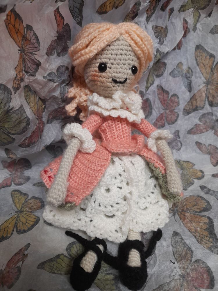 A crochet doll sitting against a paper background with butterfly patterns. She has light pink hair tied into a ponytail. She wears a dark pink blouse with white ruffles, and a layered skirt of pink and green, with white lace underneath. On her feet are black ballerina shoes. 