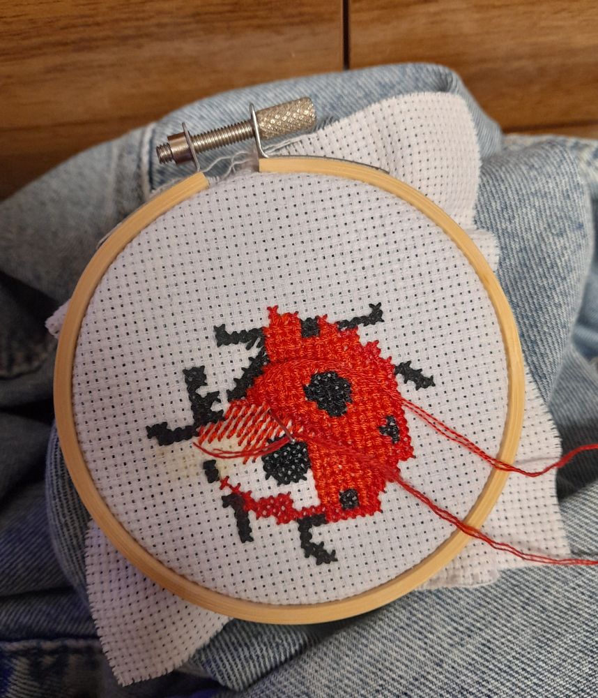 A ladybug cross stitch sitting in an embroidery hoop on a denim jacket. It is in the process of being completed, with half of the body having either a half stitch or a blank white space where stitches should be. 
